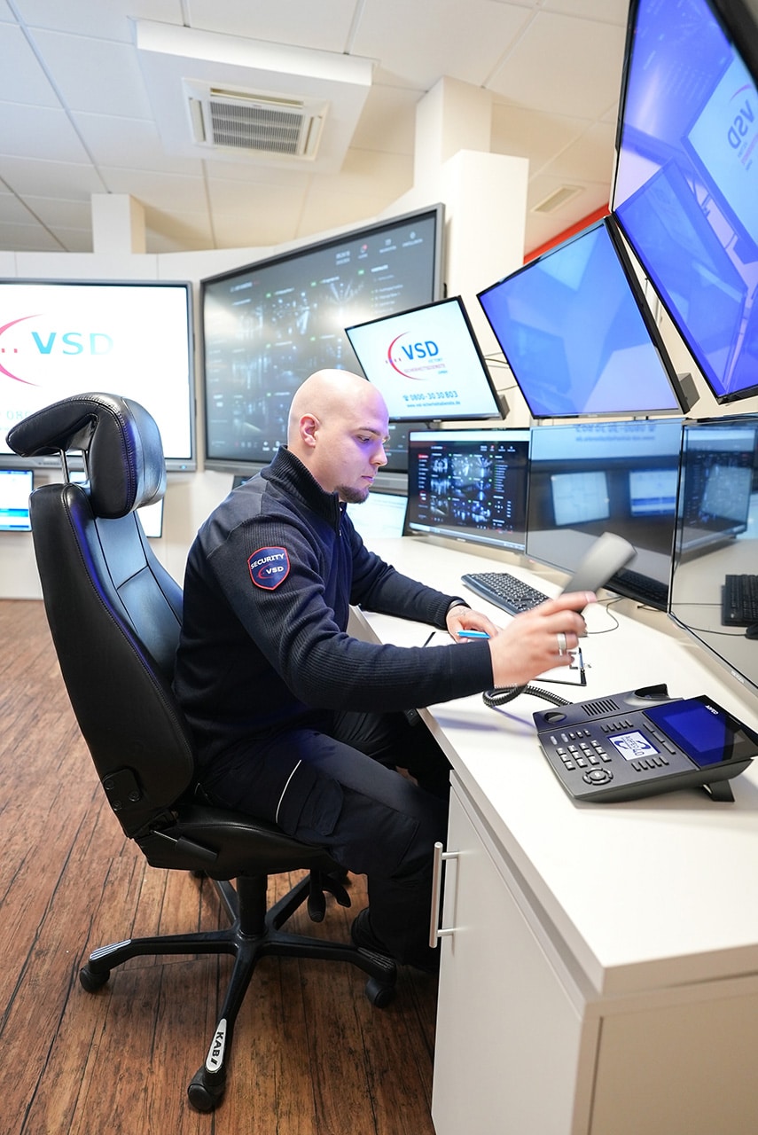 Zwei VSD Victory Sicherheitsdienste GmbH Mitarbeiter in der Leitstelle bei der Koordination eines Einsatzes – ein Mitarbeiter am Telefon, der andere zeigt auf den Überwachungsmonitor